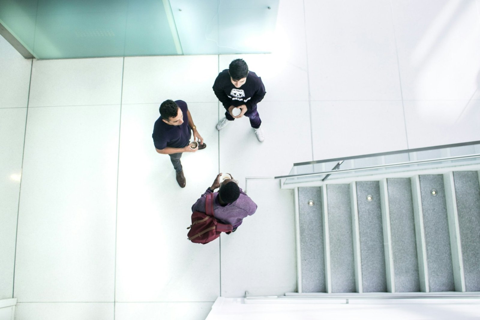 Three colleagues talking over coffee in a bright stairwell, seen from above